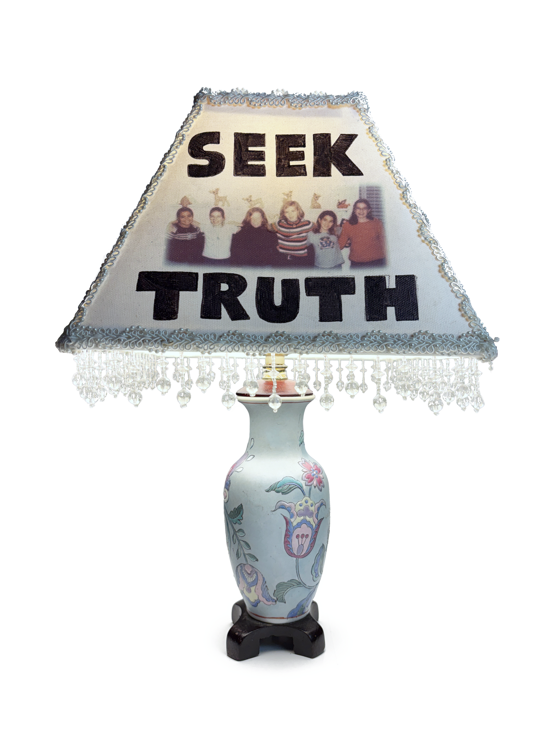 Table lamp with a white porcelain vase base painted with pastel flowers. Its beaded shade is lettered SEEK TRUTH around a photo of a group of children.